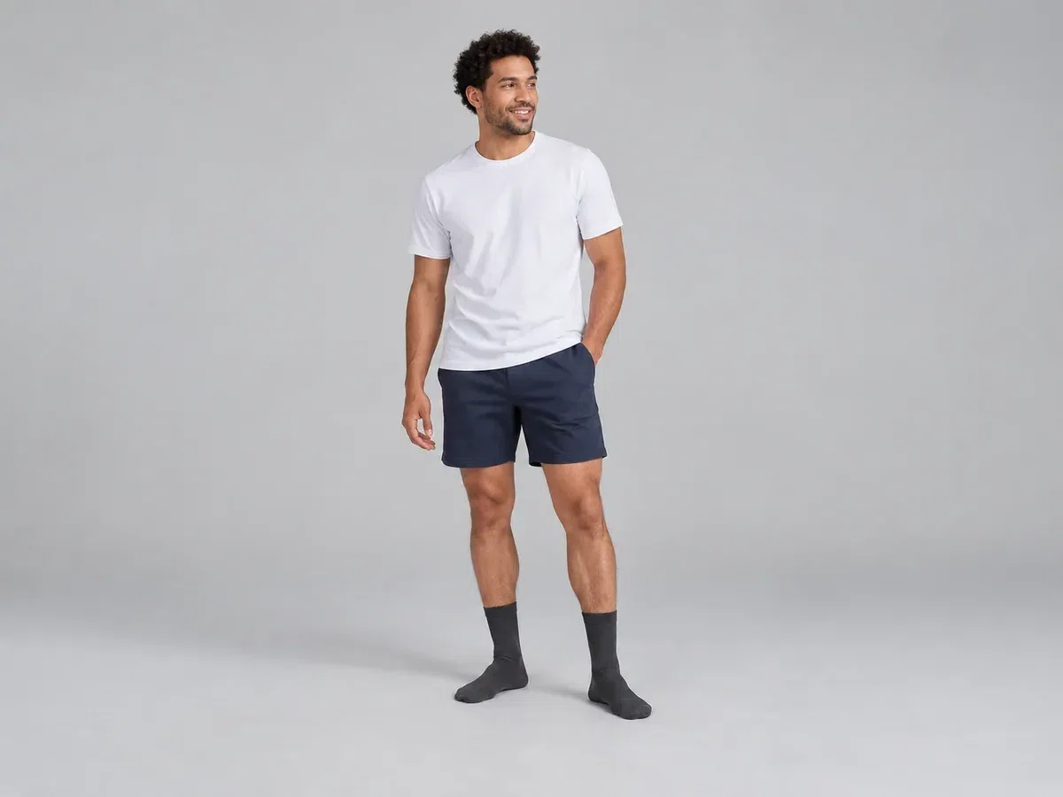 Man standing in dark socks, navy shorts and a white shirt against a grey background